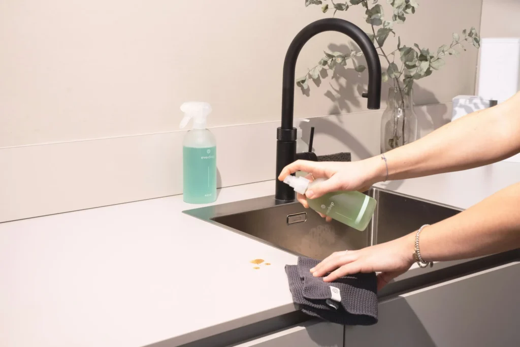 A person spraying cleaner on a kitchen counter beside the sink and wiping it with a cloth