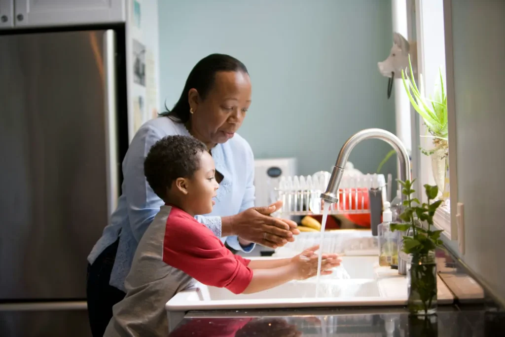 A caregiver helping a young boy wash his hands at the kitchen sink
